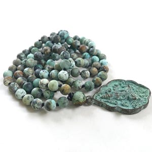 May include: A necklace featuring round, matte-finish turquoise beads with dark brown and black matrix patterns, strung on a black cord. Attached to the beads is a scalloped, shield-shaped metal pendant with a weathered, oxidized green patina finish.