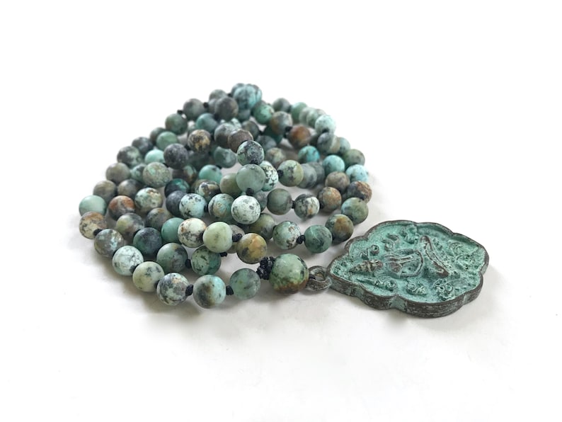 May include: A necklace featuring round, matte-finish turquoise beads with dark brown and black matrix patterns, strung on a black cord. Attached to the beads is a scalloped, shield-shaped metal pendant with a weathered, oxidized green patina finish.
