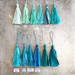 May include: Twelve fabric tassels arranged in two rows, featuring various shades of blue and green. Each tassel has a hanging loop and is labeled with names including Seafoam, Mint Julip, Shamrock, Mint, Under Water, Pacific Blue, Yosemeti Blue, Maya Blue, Deep Turquoise, Peacock Blue, Tropical Waters, and Deep Teal.