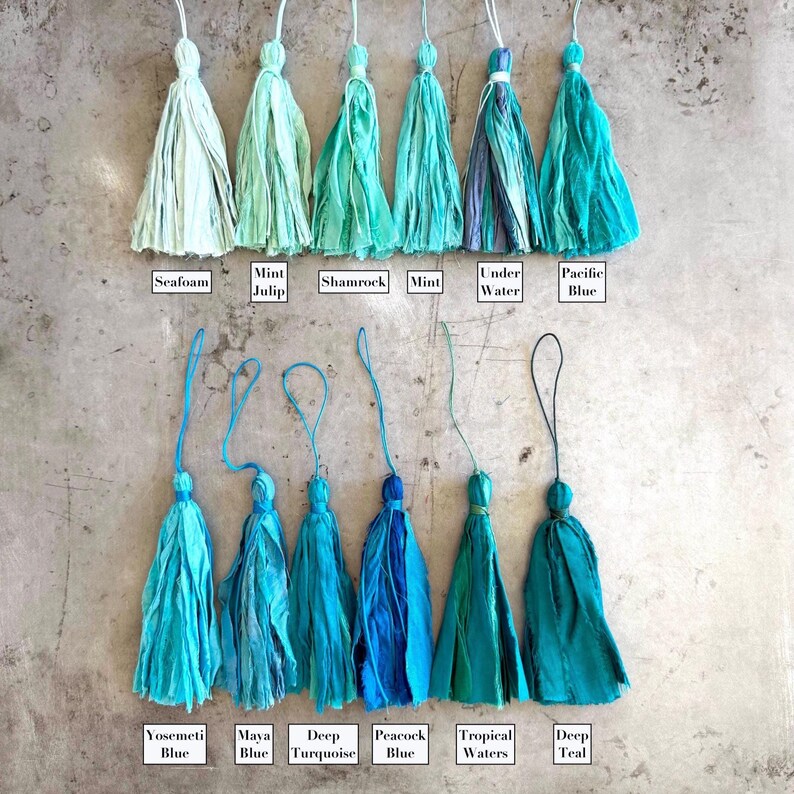 May include: Twelve fabric tassels arranged in two rows, featuring various shades of blue and green. Each tassel has a hanging loop and is labeled with names including Seafoam, Mint Julip, Shamrock, Mint, Under Water, Pacific Blue, Yosemeti Blue, Maya Blue, Deep Turquoise, Peacock Blue, Tropical Waters, and Deep Teal.