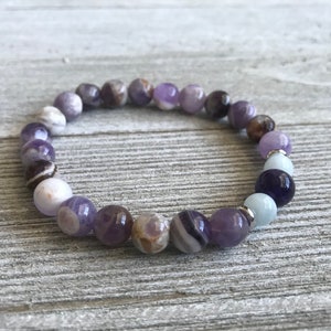 May include: A stretch bracelet composed of round beads in varying shades of purple, white, brown, and light blue. The beads feature natural marbling and banding patterns, interspersed with small silver-toned metal spacers throughout the circular design.