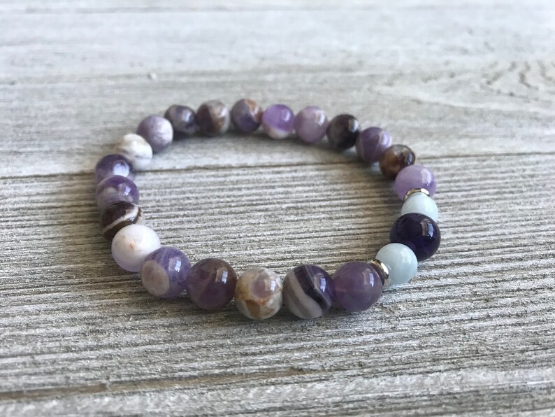 May include: A stretch bracelet composed of round beads in varying shades of purple, white, brown, and light blue. The beads feature natural marbling and banding patterns, interspersed with small silver-toned metal spacers throughout the circular design.