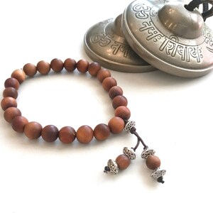 May include: A brown wooden bead bracelet featuring smooth, round beads strung on a cord. The bracelet includes two dangling ends, each finished with a single wooden bead and decorative silver-toned metal spacers. Behind it sit two brass cymbals.