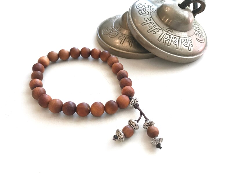 May include: A brown wooden bead bracelet featuring smooth, round beads strung on a cord. The bracelet includes two dangling ends, each finished with a single wooden bead and decorative silver-toned metal spacers. Behind it sit two brass cymbals.