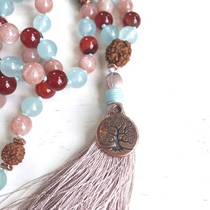 Puede incluir: Un collar de mala o cuentas de oraci&oacute;n con un dije de &aacute;rbol de la vida y una borla. El collar est&aacute; hecho con cuentas de color azul claro, rosa y rojo.
