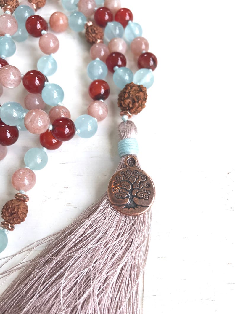 Puede incluir: Un collar de mala o cuentas de oraci&oacute;n con un dije de &aacute;rbol de la vida y una borla. El collar est&aacute; hecho con cuentas de color azul claro, rosa y rojo.