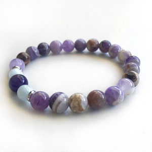 May include: A beaded bracelet featuring a variety of round, polished stones in shades of purple, lavender, brown, and light blue. The bracelet includes small silver-toned metal spacers between some of the beads, creating a multi-colored pattern.