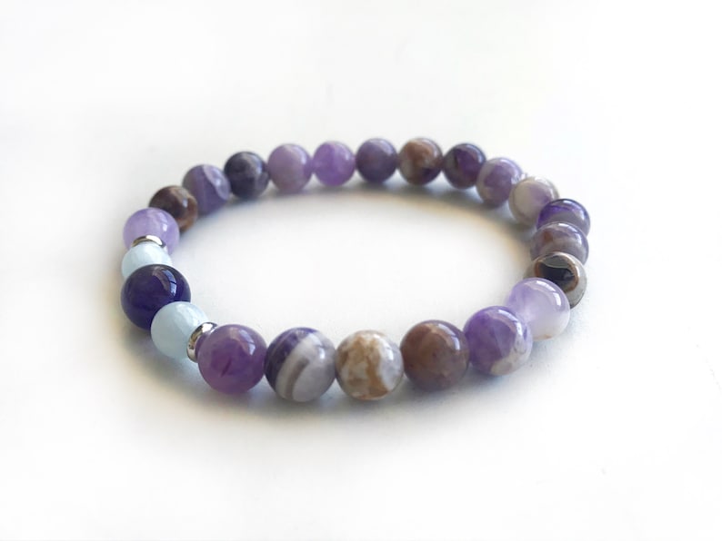 May include: A beaded bracelet featuring a variety of round, polished stones in shades of purple, lavender, brown, and light blue. The bracelet includes small silver-toned metal spacers between some of the beads, creating a multi-colored pattern.