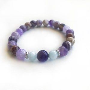 May include: A stretch bracelet composed of round beads in shades of purple, lavender, brown, and light blue. The design features polished amethyst-toned stones, earthy brown patterned beads, and two smooth light blue beads flanking a dark purple center bead.