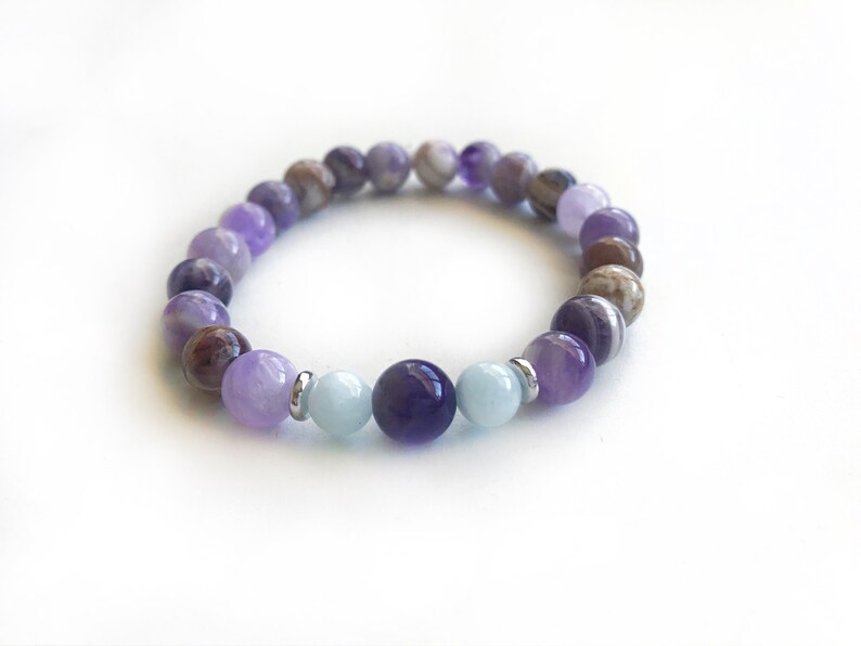 May include: A stretch bracelet composed of round beads in shades of purple, lavender, brown, and light blue. The design features polished amethyst-toned stones, earthy brown patterned beads, and two smooth light blue beads flanking a dark purple center bead.