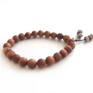 May include: A bracelet composed of round, matte brown wooden beads strung on a cord. The bracelet features a sliding knot closure with two dangling cord ends, each finished with a small, textured silver-toned metal bead and a single wooden bead.