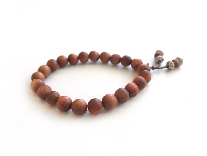 May include: A bracelet composed of round, matte brown wooden beads strung on a cord. The bracelet features a sliding knot closure with two dangling cord ends, each finished with a small, textured silver-toned metal bead and a single wooden bead.