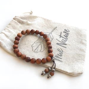 May include: A brown wooden bead bracelet with a matte finish, featuring two dangling beads accented with silver-toned metal caps. The bracelet rests on a beige drawstring pouch made of fabric, which displays a printed feather graphic and the text True Nature.