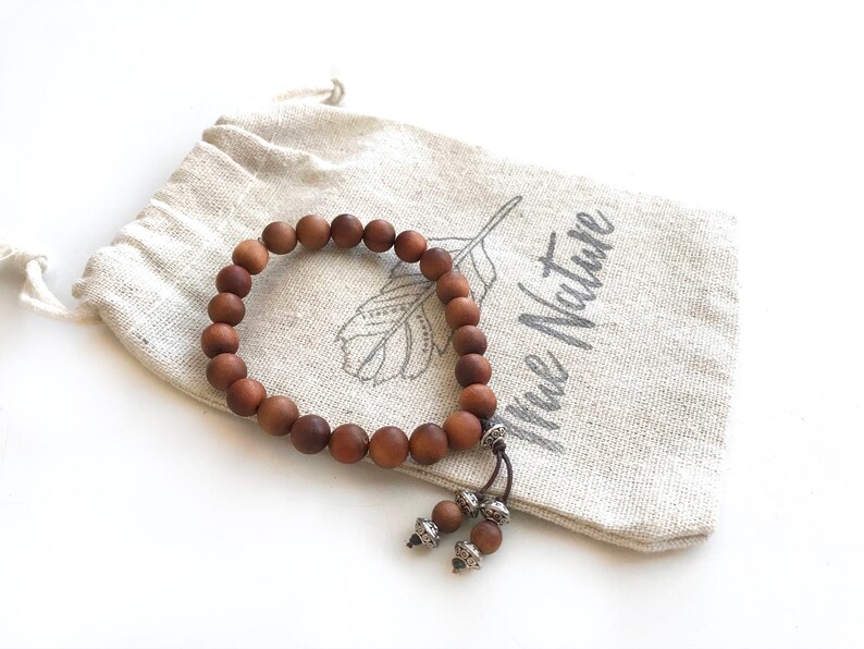 May include: A brown wooden bead bracelet with a matte finish, featuring two dangling beads accented with silver-toned metal caps. The bracelet rests on a beige drawstring pouch made of fabric, which displays a printed feather graphic and the text True Nature.
