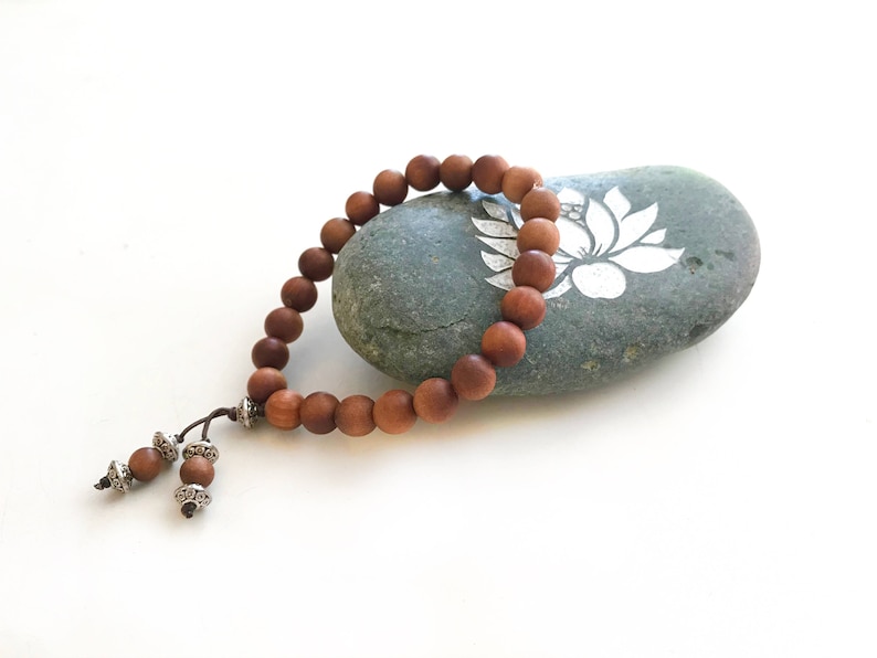 May include: A bracelet composed of smooth, round, medium-brown wooden beads strung on a dark cord. The bracelet features two dangling ends, each finished with a smaller wooden bead and a decorative, intricate silver-toned metal spacer bead.