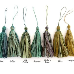 May include: Seven fabric tassels in various shades of green, ranging from dark teal to olive and mustard yellow. Each tassel features frayed edges, a tied top, and a thin hanging loop. Labels below read: Juniper, India, Dill Pickle, Chateau, Military Green, Olive, Oregano.