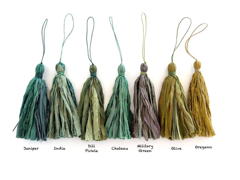 May include: Seven fabric tassels in various shades of green, ranging from dark teal to olive and mustard yellow. Each tassel features frayed edges, a tied top, and a thin hanging loop. Labels below read: Juniper, India, Dill Pickle, Chateau, Military Green, Olive, Oregano.