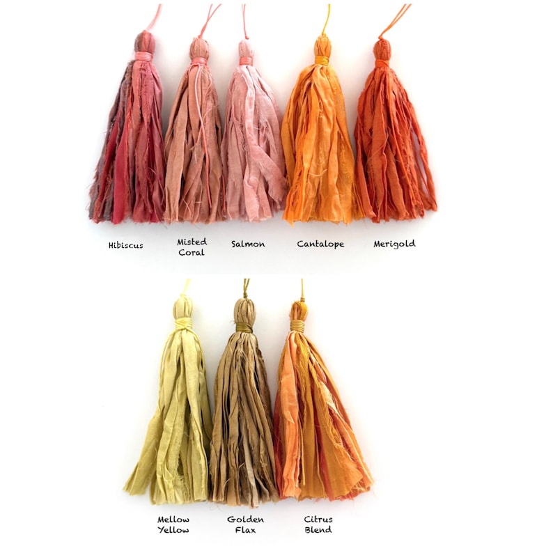 May include: A collection of eight fabric tassels arranged in two rows. The top row features Hibiscus, Misted Coral, Salmon, Cantalope, and Merigold colors. The bottom row displays Mellow Yellow, Golden Flax, and Citrus Blend. Each tassel has a tied top.