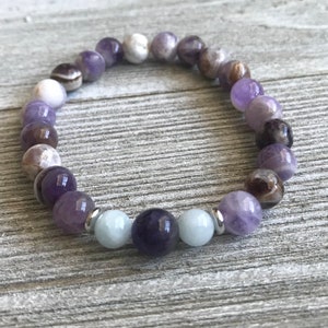 May include: A beaded bracelet featuring a circular arrangement of polished spherical stones in shades of purple, white, and dark brown, accented by two light blue beads and small silver-toned metal spacers positioned near the center of the strand.