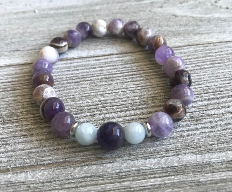 May include: A beaded bracelet featuring a circular arrangement of polished spherical stones in shades of purple, white, and dark brown, accented by two light blue beads and small silver-toned metal spacers positioned near the center of the strand.