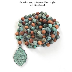 May include: A mala necklace featuring alternating round turquoise-colored stone beads and smooth brown sandalwood beads, strung on a dark cord. A large, ornate, oxidized green metal pendant with an embossed figure hangs from the bottom of the strand.
