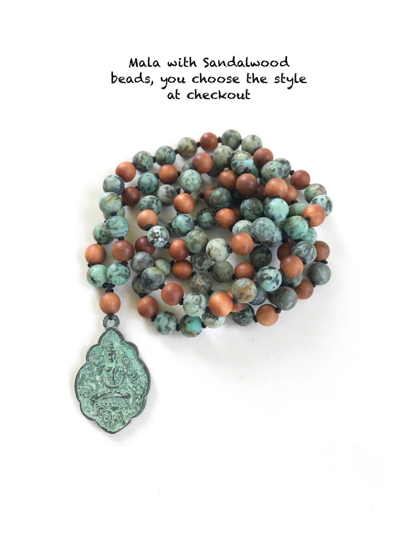 May include: A mala necklace featuring alternating round turquoise-colored stone beads and smooth brown sandalwood beads, strung on a dark cord. A large, ornate, oxidized green metal pendant with an embossed figure hangs from the bottom of the strand.