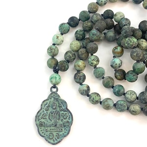 May include: A necklace featuring round, mottled green and brown stone beads strung on a dark cord. A decorative, shield-shaped metal pendant with a weathered patina hangs from the strand, displaying a central figure and intricate embossed markings.