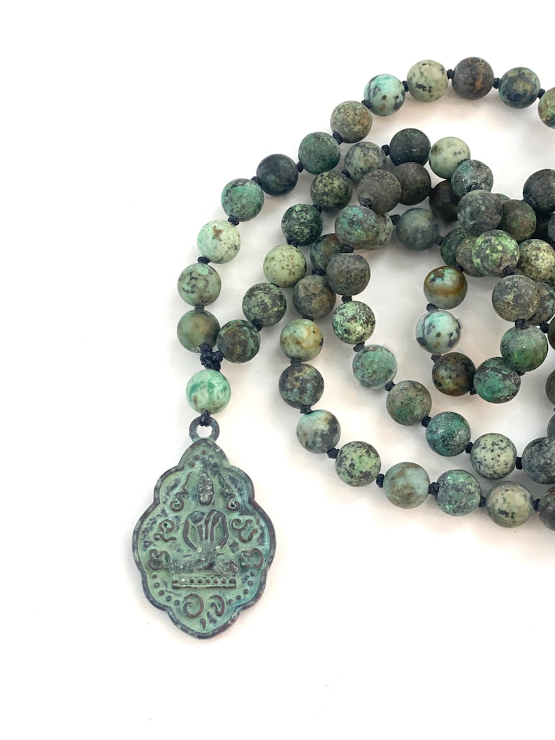 May include: A necklace featuring round, mottled green and brown stone beads strung on a dark cord. A decorative, shield-shaped metal pendant with a weathered patina hangs from the strand, displaying a central figure and intricate embossed markings.