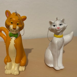 May include: Two cat figurines, one orange with a green bow tie and the other white with a yellow collar. Both are Christmas ornaments.