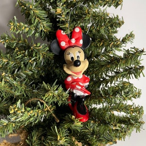 Puede incluir: Un adorno navideño de Minnie Mouse de lunares rojos y blancos. El adorno está hecho de plástico y tiene una cuerda para colgar.