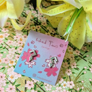 May include: A pair of silver earrings with a geometric design. The earrings are on a pink and blue card with the text "Inked Paws Earrings" and a paw print.