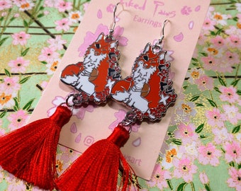 May include: A pair of dangle earrings featuring a fox design with red, white, and black accents. The earrings have red tassels and are displayed on a pink card with the text "Paws Earrings". The background is a floral pattern.
