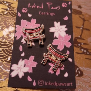 May include: A pair of gold earrings featuring a red and black Japanese torii gate design. The earrings are on a black card with the text "Inked Paws Earrings" and a social media handle "@inkedpawsart".