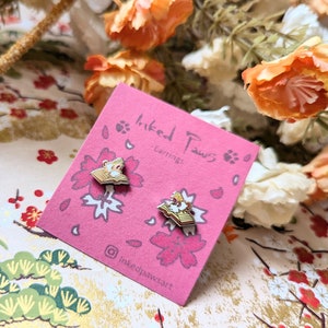 May include: A pair of gold-toned earrings featuring a small, detailed illustration of a cat reading a book. The earrings are on a pink card with the text "inked paws earrings" and a social media handle.