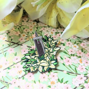 May include: An embroidered patch depicting a brown building with a sword protruding from the top, surrounded by white flowers and green leaves.