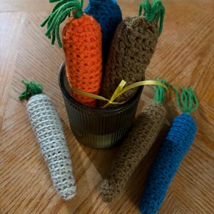 May include: A collection of handmade crochet carrots in various colors, including orange, blue, brown, and gray, with green yarn tops. Some carrots are in a dark glass container, others are arranged around it. A yellow ribbon is tied around the container.