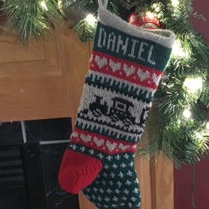 May include: A knitted Christmas stocking with a green, white, and red pattern. The stocking has the name "DANIEL" knitted into the top and a train design in the middle. The stocking is hanging on a wooden mantle.