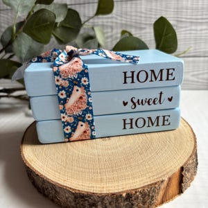May include: Three light blue wooden blocks stacked together, inscribed with the words "HOME sweet HOME". A decorative ribbon featuring a floral and hedgehog design is wrapped around the blocks. The blocks are presented on a wooden slice.