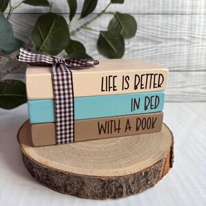 May include: Three painted wooden book blocks in beige, teal, and brown, with the text "LIFE IS BETTER IN BED WITH A BOOK". A brown and white checked ribbon binds the blocks, which are displayed on a wooden slice, ideal for a bookshelf.