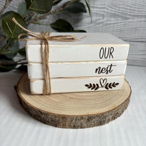 May include: A stack of three white wooden blocks with the words "OUR nest" and a small heart with leaves on the bottom block. The blocks are tied together with twine.