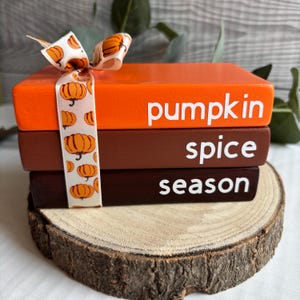 May include: Three decorative wooden books in orange and brown, with the words "pumpkin", "spice", and "season" in white. A ribbon with pumpkin designs is tied around the books, which are displayed on a wooden slice.