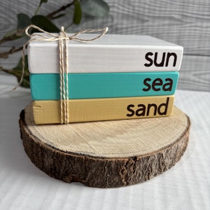 May include: Three painted wooden blocks, white, teal, and yellow, with the words "sun", "sea", and "sand" in black. The blocks are tied with twine and rest on a wooden slice, evoking a coastal theme.