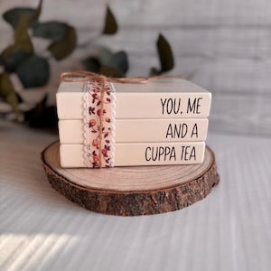 May include: Three off-white wooden blocks stacked, with the text "YOU. ME AND A CUPPA TEA" in black. A floral ribbon and twine bind the blocks, which sit on a wooden slice.