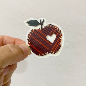 May include: A red apple sticker made of pencils with a heart shape cut out of the center. The apple has a green stem and leaf.