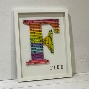 May include: A white framed picture with a rainbow letter F made of crayons. The name Finn is written in black below the letter.