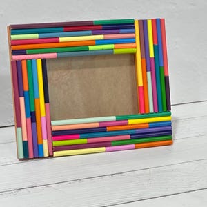 May include: A rectangular photo frame with a brown interior and a colorful exterior. The frame is decorated with an array of small, brightly colored sticks in shades of pink, yellow, green, blue, and orange.