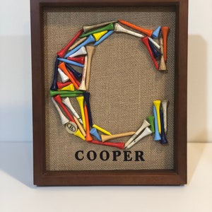 May include: A framed burlap canvas with the letter "C" made from colorful golf tees. The name "COOPER" is printed below the letter.