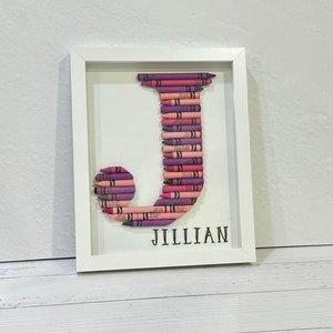 May include: A white framed picture with a letter J made out of pink and purple crayons. The name Jillian is written below the letter.