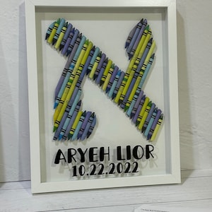 May include: A white framed picture with a Hebrew letter Aleph made from colorful crayons. The letter is surrounded by a white background with the name "Aryeh Lior" and the date "10.22.2022" written in black.