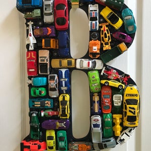 May include: A black letter B made of wood with a variety of toy cars glued to the surface. The cars are in various colors, including red, blue, green, yellow, and orange.